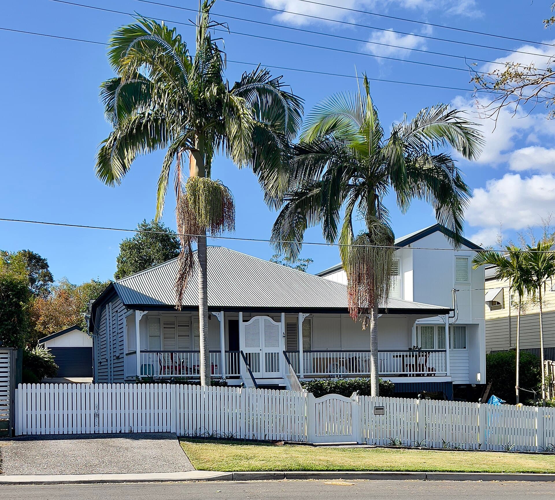 White weatherboard Queenslander with a corrugated-iron roof, verandah and picket fence, flanked by two palm trees