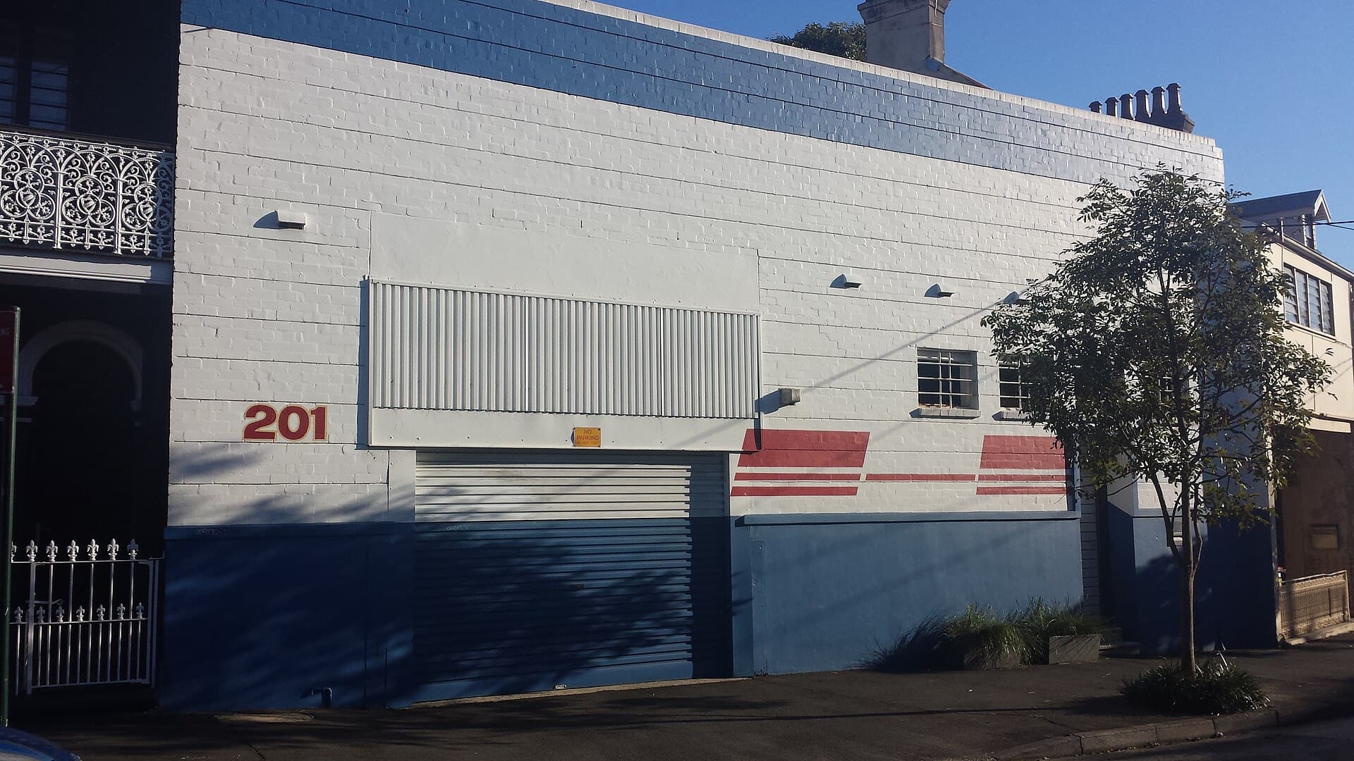 White and blue painted warehouse facade with a roller door and the street number 201 on a quiet suburban street