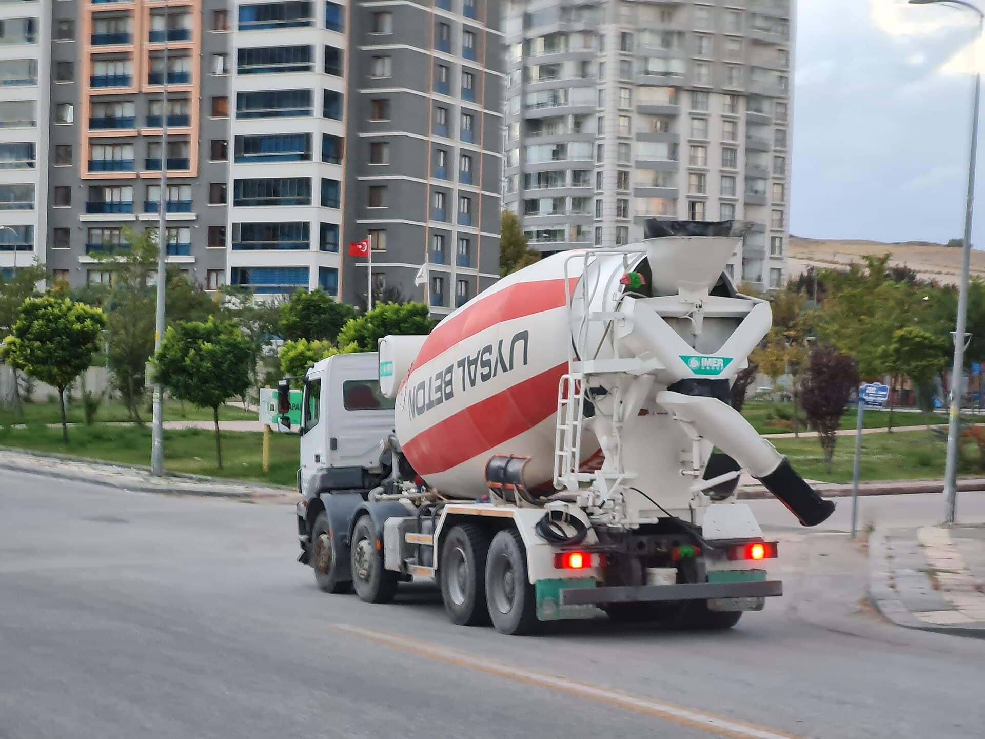 White concrete mixer truck with a red stripe driving along a suburban road, apartment towers in the background