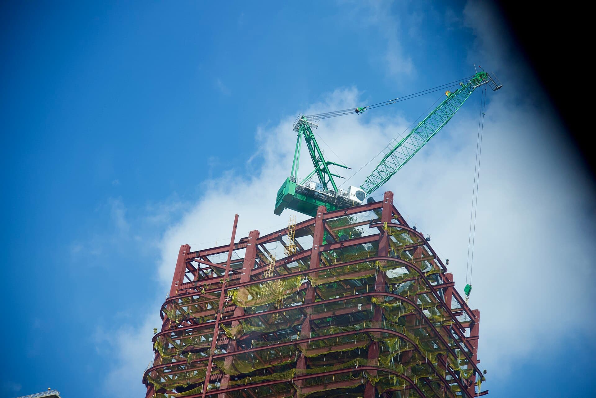 Red steel frame of a mid-rise building under construction, wrapped in yellow safety mesh, with a green tower crane against a blue sky
