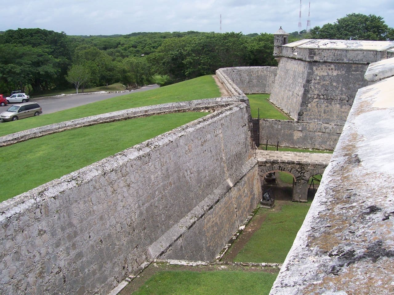 Thick pale masonry fortress walls meeting at a corner, with a grassed courtyard and an arched passage below